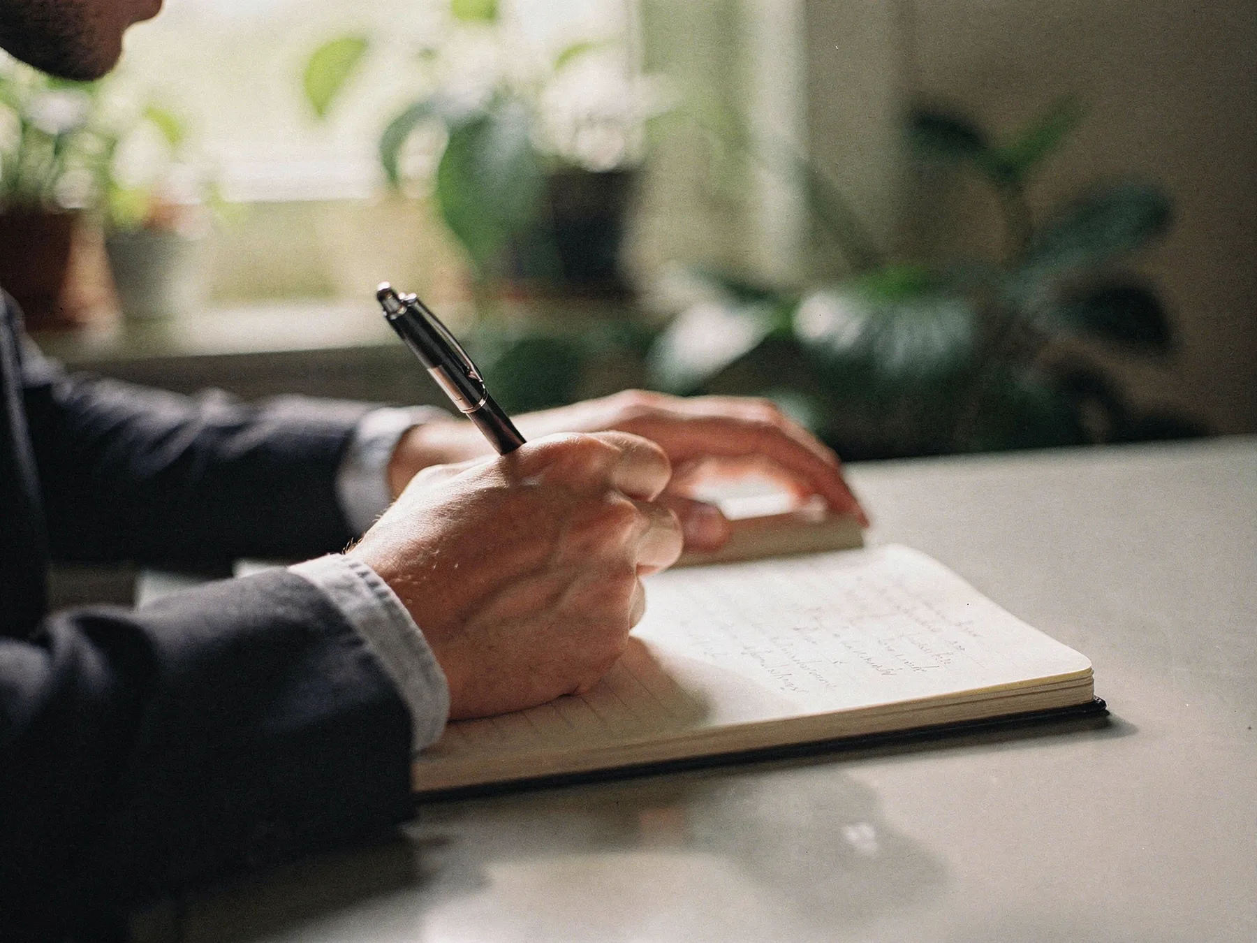 Hände schreiben mit einem Stift in ein Notizbuch, im Hintergrund grüne Pflanzen